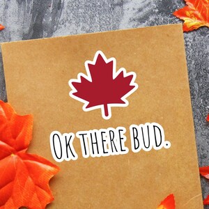 Peut inclure: Une carte brune avec une feuille d'érable rouge et le texte "OK THERE BUD." entourée de feuilles d'automne orange et jaunes.