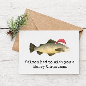 May include: A Christmas card featuring a watercolor illustration of a salmon wearing a Santa hat. The card reads "Salmon had to wish you a Merry Christmas." The card is on a white wooden surface with a brown envelope and pine sprig.