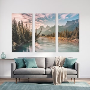 Può includere: Un set di stampe su tela a trittico raffigurante una serena scena fluviale con montagne e alberi sempreverdi. L'opera presenta un fiume verde acqua, un cielo nuvoloso e una tavolozza di colori neutri. Il set è esposto sopra un divano grigio con cuscini verde acqua.