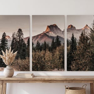 Peut inclure: Impression sur toile en trois parties représentant un paysage de montagne avec une forêt au premier plan. L'œuvre utilise une palette de couleurs chaudes avec des tons de marron, beige et vert. Un vase avec de l'herbe de la pampa séchée est posé sur une table en bois.