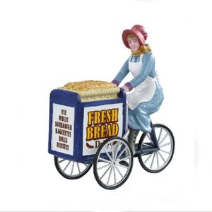Op de afbeelding: Een miniatuurbeeldje van een vrouw op een fiets met een broodkar. De kar is blauw en wit met "FRESH BREAD" in gouden letters. De kar vermeldt ook "Rye, Wheat, Sourdough, Baguettes, Rolls, Biscuits". De vrouw draagt een blauwe jurk en een roze hoed.