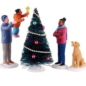 Könnte beinhalten: Eine Familie schmückt einen kleinen grünen Weihnachtsbaum mit goldenem Lametta und bunten Kugeln. Ein Kind setzt einen Stern auf die Spitze des Baumes. Ein Hund beobachtet die Familie.