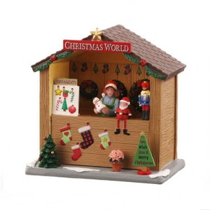 Op de afbeelding: Een houten kerstmarktkraam met een bruin dak en een bord met de tekst "Christmas World". De kraam is versierd met kerstversiering, kousen en een kleine kerstboom. Er zijn ook poppetjes van mensen en een notenkraker.