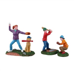 May include: A baseball-themed figurine set featuring a girl pitching, a dog with a bat, and two boys playing baseball. The figures are painted with vibrant colors, including blue, red, and yellow, and are set on green bases.