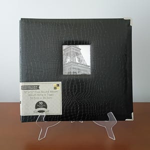 May include: Black 12" x 12" post-bound photo album with a textured cover and a small square window displaying a black and white image of the Eiffel Tower. The album has metal corner protectors and a label indicating it holds 10 top-loading pages.
