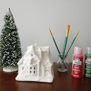 May include: A white ceramic house figurine, a miniature green pine tree with white snow-like flocking, three paintbrushes in a small glass vase, and two 2 fl oz bottles of FolkArt Matte acrylic paint in red and green colors.