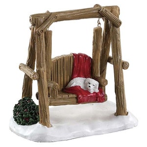 May include: A white dog figurine is sleeping on a red blanket in a wooden swing. The swing is made of rough-hewn logs and has a rustic look. The dog is lying on its side with its head resting on the blanket. The swing is set against a snowy background.