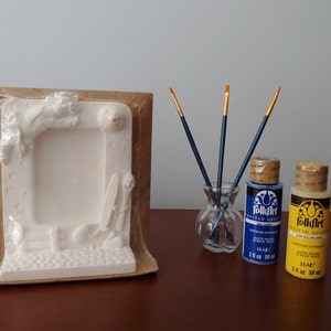 May include: A white ceramic picture frame with a winter scene design, packaged in plastic. Two bottles of FolkArt acrylic paint, one blue and one yellow, are next to a glass vase with three paintbrushes.