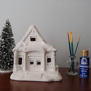 May include: A white ceramic general store building with a snow-covered roof, ready to be painted. The building has a sign that reads "General Store".  Two bottles of acrylic paint, one blue and one red, are next to a glass jar with three paintbrushes.