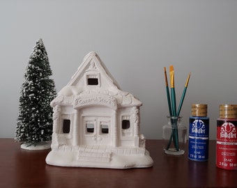 May include: A white ceramic general store building with a snow-covered roof, ready to be painted. The building has a sign that reads "General Store".  Two bottles of acrylic paint, one blue and one red, are next to a glass jar with three paintbrushes.
