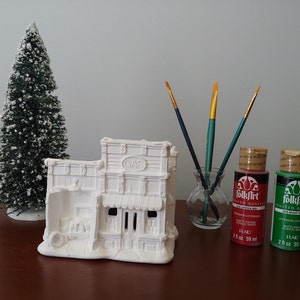 May include: A white ceramic building with the word "GAS" on the front, a small green Christmas tree, paintbrushes, and two bottles of FolkArt acrylic paint. The paint bottles are red and green, each containing 2 fl oz.