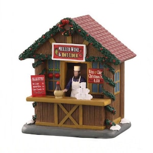 May include: A miniature wooden Christmas market stall with a red roof and green garland. The stall has a sign that reads "Mulled Wine & Hot Cider". A figure wearing a white apron stands behind the counter.