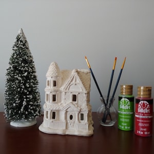 May include: A white ceramic house with a snow-covered pine tree, two paintbrushes in a glass vase, and two bottles of FolkArt acrylic paint in green and red.
