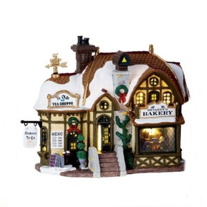 May include: A miniature Christmas village building with a snow-covered roof. The building has signs that read "Ye Olde Tea Shoppe" and "Devaney's Bakery". It features a chimney and a sign that says "Orders To Go".