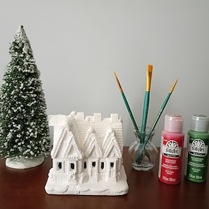 May include: A white ceramic miniature building with three gabled roofs, a small artificial pine tree with snow-dusted branches, three paintbrushes in a glass jar, and two 2 fl oz bottles of FolkArt matte acrylic paint in red and green colors.