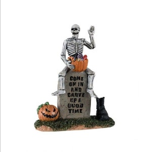 Peut inclure: Une figurine de squelette assise sur une pierre tombale avec le texte "Come on in and carve up a good time". Le squelette tient une citrouille remplie de bonbons. Il y a une petite citrouille devant la pierre tombale.