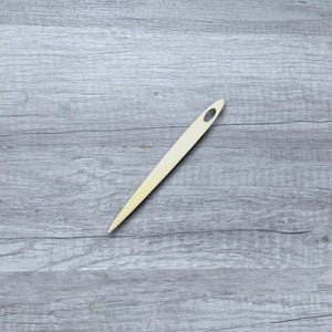 May include: A long, thin, wooden needle with a small eye at the top. The needle is lying on a light gray wooden surface.