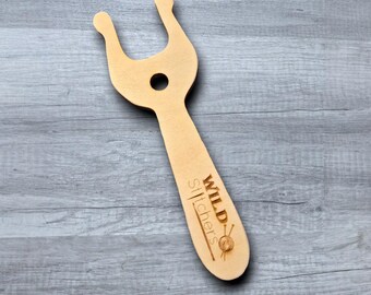 May include: A light-colored wooden yarn swift with a handle and a circular hole. The top has a U-shaped design with two rounded ends. The handle has the text "WILD Stitchers" and a small yarn ball graphic.
