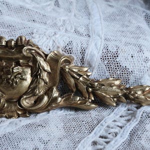 Op de afbeelding: Een goudkleurige metalen decoratieve trim met een bloemmotief. De trim heeft een centraal schildvormig motief met ingewikkelde details en een bladachtig, rolvormig ontwerp dat zich naar rechts uitstrekt.