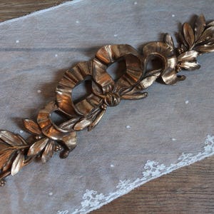 May include: A metallic bronze-toned decorative applique featuring a central ribbon bow motif flanked by symmetrical leafy laurel branches. The elongated ornament displays a sculpted, three-dimensional relief design with a polished finish.