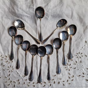 Puede incluir: Vista aérea de una colección de cucharas de plata antiguas dispuestas sobre un paño blanco bordado. Las cucharas, de varios tamaños, tienen cuencos redondeados y mangos largos y delgados. El paño presenta un delicado motivo floral.