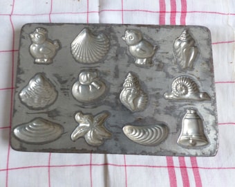 Op de afbeelding: Een vintage metalen mal voor het maken van chocolade snoepjes. De mal heeft 12 verschillende vormen, waaronder schelpen, een slak, een bel en een zeester.