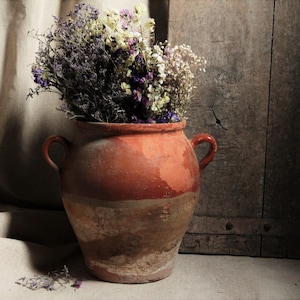以下が含まれることがあります： 紫、白、黄色のドライフラワーが入った、取っ手付きの素朴なテラコッタ色の花瓶。 花瓶はニュートラルな色の表面に置かれ、背景には木のパネルがあります。 全体的な雰囲気はヴィンテージで自然です。