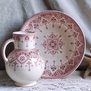 Könnte beinhalten: Ein weißes Keramik-Krug- und Teller-Set mit einem bordeauxfarbenen Blumenmuster. Der Krug hat einen runden Körper und einen Henkel. Der Teller ist groß und rund. Ein Seestern und eine Muschel befinden sich im Hintergrund.