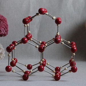 May include: A molecular model structure composed of numerous glossy red spherical atoms connected by thin silver metallic rods, forming a repeating hexagonal lattice pattern. The structure is displayed as a three-dimensional geometric arrangement.