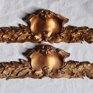 May include: Two identical gold-colored decorative architectural pediments featuring a central shell motif flanked by symmetrical carved leaf and floral garlands. These elongated, horizontal ornaments display a metallic finish with intricate relief details.