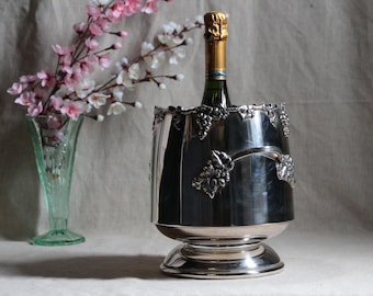 May include: Silver wine cooler with a grapevine design and a bottle of champagne inside. The cooler has a round base and a wide, cylindrical body. The grapevine design is embossed on the cooler and features leaves and grapes.