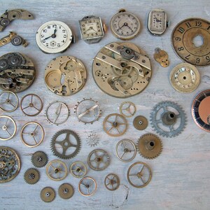 May include: A collection of vintage watch parts and gears, including clock faces, hands, and mechanisms. The parts are arranged on a light wood surface.