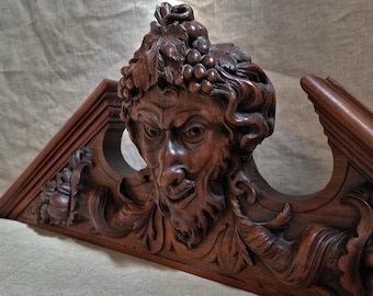 May include: A detailed, dark brown wooden carving of a face with a beard and grape leaves in its hair. The carving is part of a larger, ornate wooden structure with triangular elements and floral details.