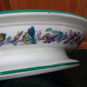 May include: A white ceramic pedestal bowl with a green rim and floral design. The bowl has a raised base and is decorated with a delicate floral pattern in shades of pink, blue, green, and yellow.