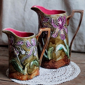 May include: Two ceramic jugs featuring a raised relief design of purple iris flowers on a textured olive green background. Each jug has a bright pink interior, a curved handle with a dotted pattern, and a decorative base with an embossed border.