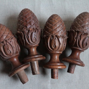 May include: Four brown wooden finials shaped like pinecones, each featuring a carved scale pattern on the top section and decorative leaf-like carvings around the base, with a cylindrical peg extending from the bottom of each piece.