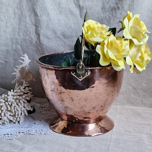 Può includere: Un secchio di rame martellato con manico, contenente un bouquet di rose gialle. Il secchio ha un interno scuro e poggia su una piccola base svasata. Una decorazione bianca simile al corallo e un panno di pizzo bianco sono sullo sfondo.