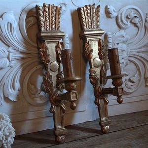 Pode incluir: Um par de arandelas de parede douradas com entalhes verticais em forma de flecha no topo, detalhes ornamentados de folhas ao longo da haste central e um suporte de vela cilíndrico que se estende pela frente com um acabamento desgastado.