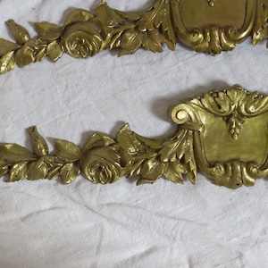 Op de afbeelding: Twee sierlijke, goudkleurige metalen decoratieve stukken met een bloemmotief. Elk stuk heeft een centraal schildvormig element met ingewikkelde krulwerk en een bloemmotief.