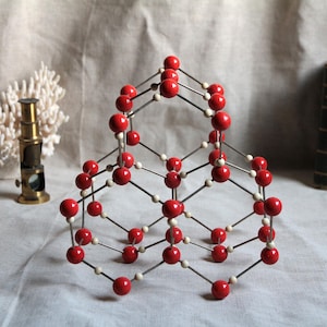 May include: A vintage scientific model with red spheres connected by silver rods, resembling a molecular structure. A small brass microscope, white coral, and two antique books are in the background, suggesting a theme of science and history.