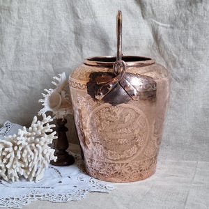 May include: A copper-colored, antique-style vase with a handle. The vase features an embossed design and is set against a neutral backdrop with decorative elements. The handle is attached with a metal clasp.