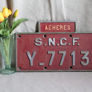 Pode incluir: Uma placa de metal vermelha vintage com o texto "ACHERES" e "S.N.C.F. Y-7713" em letras prateadas. Um vaso de vidro verde contém tulipas amarelas. A placa tem uma superfície texturizada, possivelmente um artefato ferroviário.