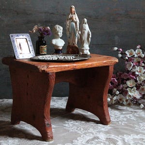 Peut inclure: Un petit tabouret rectangulaire en bois avec une finition brun chaud et quatre pieds robustes. Sur le dessus repose un plateau en métal argenté contenant un petit cadre photo, une bouteille en verre sombre avec des fleurs séchées, un buste blanc et deux figurines religieuses.
