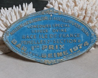 Puede incluir: Una placa ovalada de metal azul y dorado con el texto "MINISTEREDELAGRICULTURELY DUDEVELOPPEMENT RURAL CONCOURS SPECIAL ESPECE OVINE RACE ILE DE FRANCE FEMELLES 7° SECTIONB BAR SUR SEINE 1974"