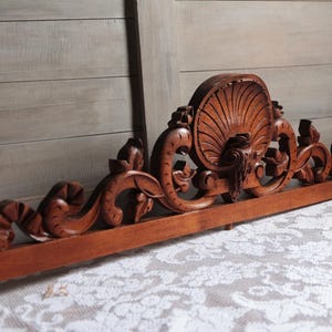 Może przedstawiać: Drewniana główka łóżka z rzeźbionymi, skomplikowanymi detalami, z motywem muszli w środku i zwiniętymi motywami kwiatowymi. Główka łóżka jest w kolorze ciemnobrązowym i ma postarzaną powierzchnię.