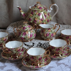 May include: A porcelain tea set featuring a teapot, a sugar bowl with a lid, a creamer, and four teacups with matching saucers. Each piece is decorated with a dense pattern of pink and yellow roses on a green background with gold trim.