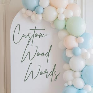 May include: A white arched sign with the words "Custom Wood Words" in green script, decorated with a pastel balloon garland in shades of blue, white, and peach. The sign is set against a white wall and wooden floor.