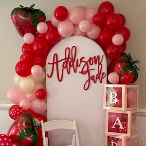 May include: A baby shower decoration featuring a white arch with the name "Addison Jade" in red script. The arch is surrounded by red, pink, and white balloons, and strawberry-shaped balloons. Wooden blocks spell "BABY". A white chair is also present.