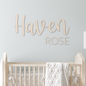 May include: A white wooden sign with the name "Haven Rose" written in cursive script. The sign is hanging on a light blue wall above a white cot.