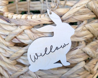 May include: Woven basket with a bunny-shaped tag that says "Willow". The basket is made of natural-colored woven material. The bunny tag is made of light-colored wood and is tied to the basket with twine. The basket has two handles.